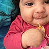 Zoé Jimena is registered to the contest to win money with this photo: baby, blue_background, cheeks, child, close_up, curly_hair, cute, expression, eyes, face, food, hand, holding, infant, lying_down, orange_fruit, pink_clothing, skin, smile, toddler
