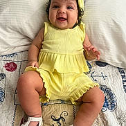 Zoé Jimena joined the competition — help win amazing prizes! baby, child, smile, yellow_clothing, headband, sitting, white_sandals, curly_hair, quilt, bed, pillow, happy, cute, indoors, infant, person, footwear, patterned_textile, playful, portrait