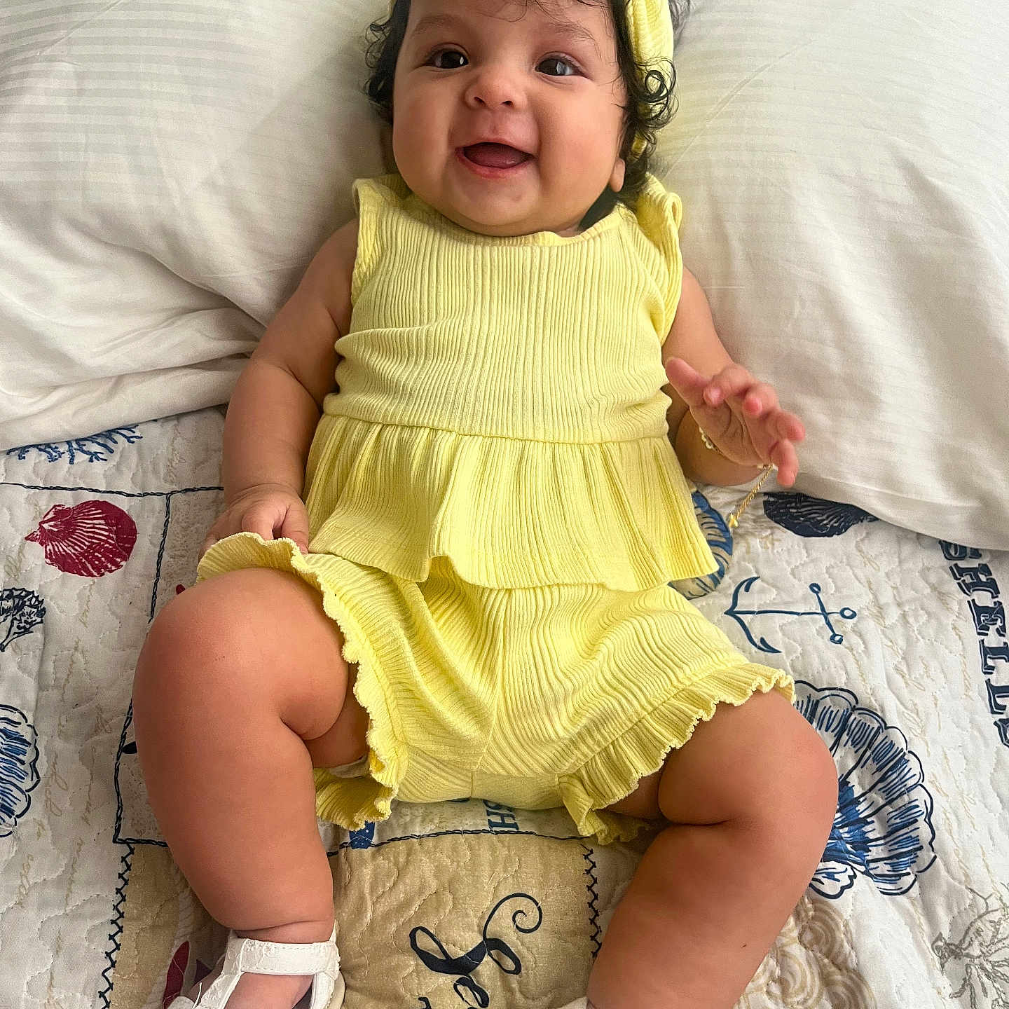 Zoé Jimena joined the competition — help win amazing prizes! baby, child, smile, yellow_clothing, headband, sitting, white_sandals, curly_hair, quilt, bed, pillow, happy, cute, indoors, infant, person, footwear, patterned_textile, playful, portrait