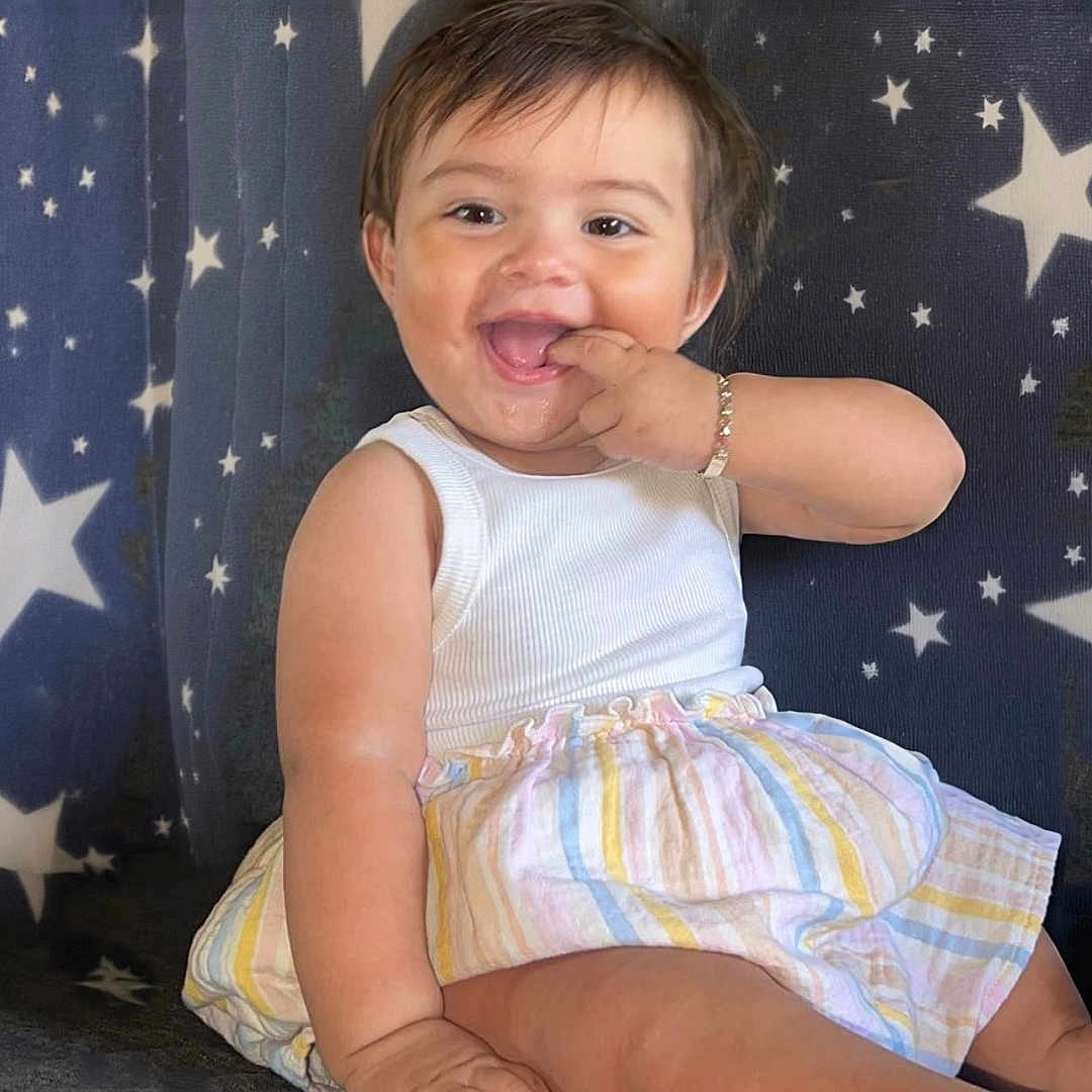 Souline is registered to the contest to win money with this photo: baby, barefoot, bracelet, child, closeup, couch, cute, fingers_in_mouth, happy, indoor, infant, joyful, playful, portrait, sitting, smiling, star_pattern, striped_skirt, tank_top, young_child