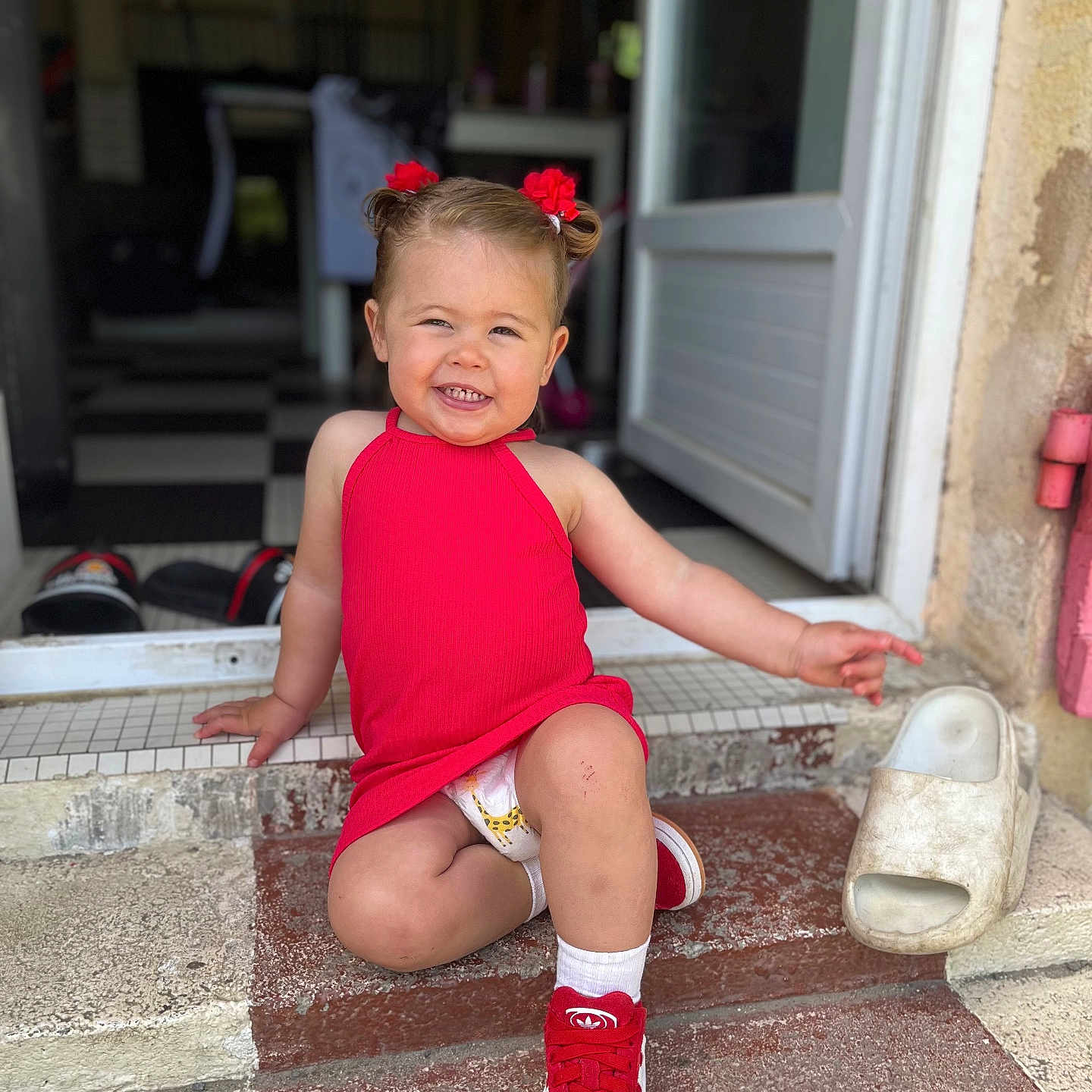 Kataleya a rejoint le concours — aidez-le/la à gagner de superbes lots ! toddler, child, smiling, red_dress, red_shoes, sitting, outdoor, steps, hair_bows, diaper, doorway, pointing, happy, casual, playful, concrete, footwear, person, cute, daylight