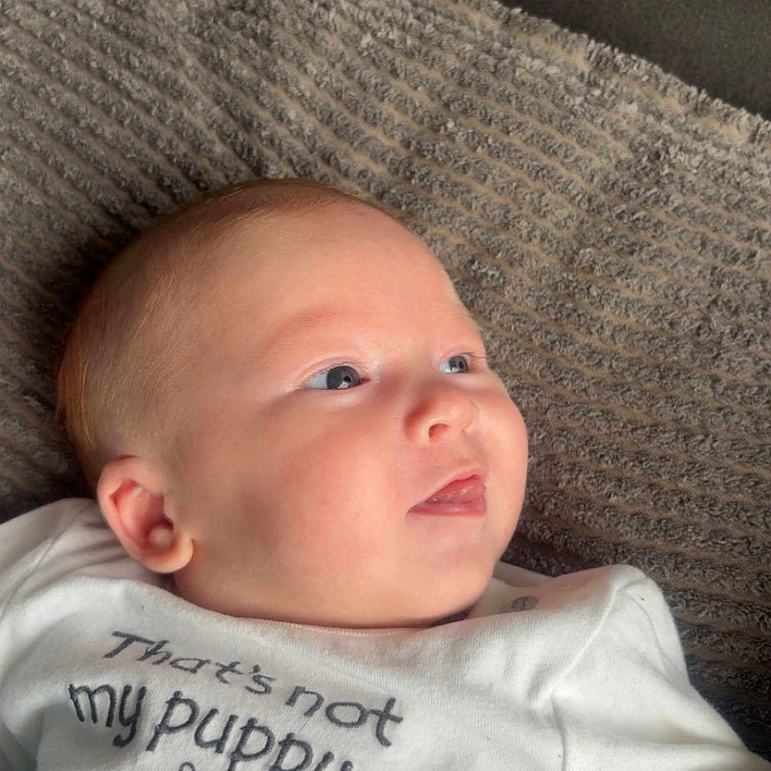 Tjay joined the competition — help win amazing prizes! baby, blanket, child, clothing, curious, cute, dog_illustration, ear, expression, face, head, indoors, infant, laying_down, person, skin, soft_lighting, text_on_clothing, texture, white_shirt