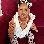 Germany is registered to the contest to win money with this photo: baby, child, clothing, couch, cozy, cute, fashion, footwear, happy, headband, indoors, leopard_print, pants, person, portrait, red_couch, sitting, smiling, toddler, vest
