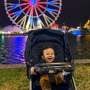 Adrian is registered to the contest to win money with this photo: toddler, child, stroller, night, ferris_wheel, carnival, lights, water, grass, smiling, happy, shoe, clothing, outdoor, amusement_park, reflection, person, baby, seat, fun