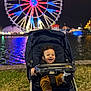 toddler, child, stroller, night, ferris_wheel, carnival, lights, water, grass, smiling, happy, shoe, clothing, outdoor, amusement_park, reflection, person, baby, seat, fun