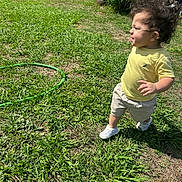 Adrian is registered to the contest to win money with this photo: toddler, child, curly_hair, yellow_shirt, beige_shorts, white_shoes, grass, sunny, outdoor, garden_hose, greenery, walking, squinting, person, background, lawn, summer, daylight, nature, yard