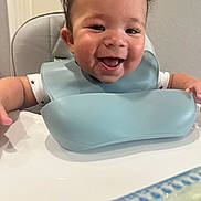 Adrian is registered to the contest to win money with this photo: arm, baby, bib, blue, child, clothing, curly_hair, cute, expression, face, feeding, happy, high_chair, indoor, infant, skin, smile, table, tongue, white