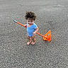toddler, child, barefoot, blue_onesie, curly_hair, orange_shovel, asphalt, outdoor, play, toy, small_person, standing, daylight, parking_lot, curious, cute, single_child, casual, summer, young_child