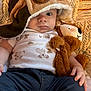 baby, child, face, aviator_hat, goggles, stuffed_animal, plush_toy, clothing, patterned_background, warning_label, hand, jeans, white_onesie, cute, indoor, portrait, infant, soft_toy, lying_down, expression