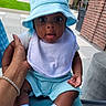 baby, infant, child, portrait, blue_hat, bucket_hat, bib, blue_clothing, sitting, held, adult_hand, outdoor, courtyard, brick_building, lawn, wide_eyes, open_mouth, baby_socks, baby_shoes, photography