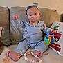 baby, infant, toddler, headband, bow, onesie, toy, rattle, teether, beaded_nursing_necklace, couch, pillow, blanket, living_room, portrait, sitting, barefoot, cute, neutral_expression, soft_lighting