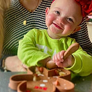 Paisley is registered to the contest to win money with this photo: baby, smiling, toddler, headband, red_bow, green_shirt, spoon, silicone_plate, food, mealtime, table, adult, striped_shirt, necklace, closeup, portrait, happy, rosy_cheeks, sitting, kitchen_background