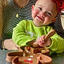 baby, smiling, toddler, headband, red_bow, green_shirt, spoon, silicone_plate, food, mealtime, table, adult, striped_shirt, necklace, closeup, portrait, happy, rosy_cheeks, sitting, kitchen_background