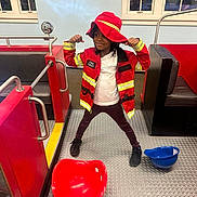 Nyla is registered to the contest to win money with this photo: child, fire_chief_costume, red_jacket, red_hat, flexing, indoor, metal_floor, plastic_seat, blue_helmet, windows, cityscape, booth_seating, black_shoes, white_shirt, play_area, confident, posing, person, young, smiling