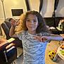 child, boy, curly_hair, tshirt, peace_sign, living_room, carpet, lamp, sofa, curtains, window, toy, soccer_ball, furniture, smile, person, indoor, casual_clothing, playful, happy