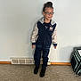 child, glasses, jacket, pants, black_shoes, carpet, indoor, wall, vent, bag, standing, person, casual_clothing, hair_bun, calm_expression, light_gray_wall, floor, young, portrait, looking_sideways