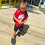 Zakari joined the competition — help win amazing prizes! child, boy, red_shirt, nike, backpack, black_shorts, black_shoes, sidewalk, steps, yellow_railings, school_building, daylight, outdoor, concrete, shadow, hair_bun, serious_expression, walking, person, casual_clothing