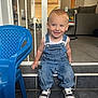 toddler, child, denim_overalls, sneakers, blue_chair, smiling, indoor, living_room, carpet, tile_floor, baby_gate, furniture, play_area, door, window, curtains, person, cute, happy, portrait