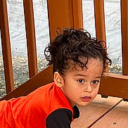 Alexander joined the competition — help win amazing prizes! child, curly_hair, orange_shirt, black_shirt, wooden_deck, wooden_railings, outdoor, crawling, young_child, person, face, curious, daylight, casual_clothing, natural_light, playful, expression, young_kid, skin_tone, hair_tied_up