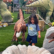 Alexander is registered to the contest to win money with this photo: adventure, child, dinosaur, dinosaur_model, dinosaur_park, faux_grass, museum, outdoor, playful, portrait, prehistoric, raptor, ropes, standing, statue, theme_park, trees, triceratops, tropical_plants, tyrannosaurus