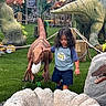 adventure, child, dinosaur, dinosaur_model, dinosaur_park, faux_grass, museum, outdoor, playful, portrait, prehistoric, raptor, ropes, standing, statue, theme_park, trees, triceratops, tropical_plants, tyrannosaurus