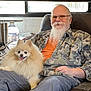 man, dog, pomeranian, beard, glasses, camouflage_shirt, orange_tshirt, indoor, window, blinds, chair, smile, happy, pet, fluffy_dog, relaxing, seated, laptop, water_bottle, cozy