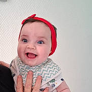 Sélène participe au concours pour gagner de l'argent avec cette photo : adult_hand, baby, bib, blue_eyes, child, clothing, cute, expression, face, hand, happy, headband, indoor, infant, person, portrait, skin, smile, texture, white_wall