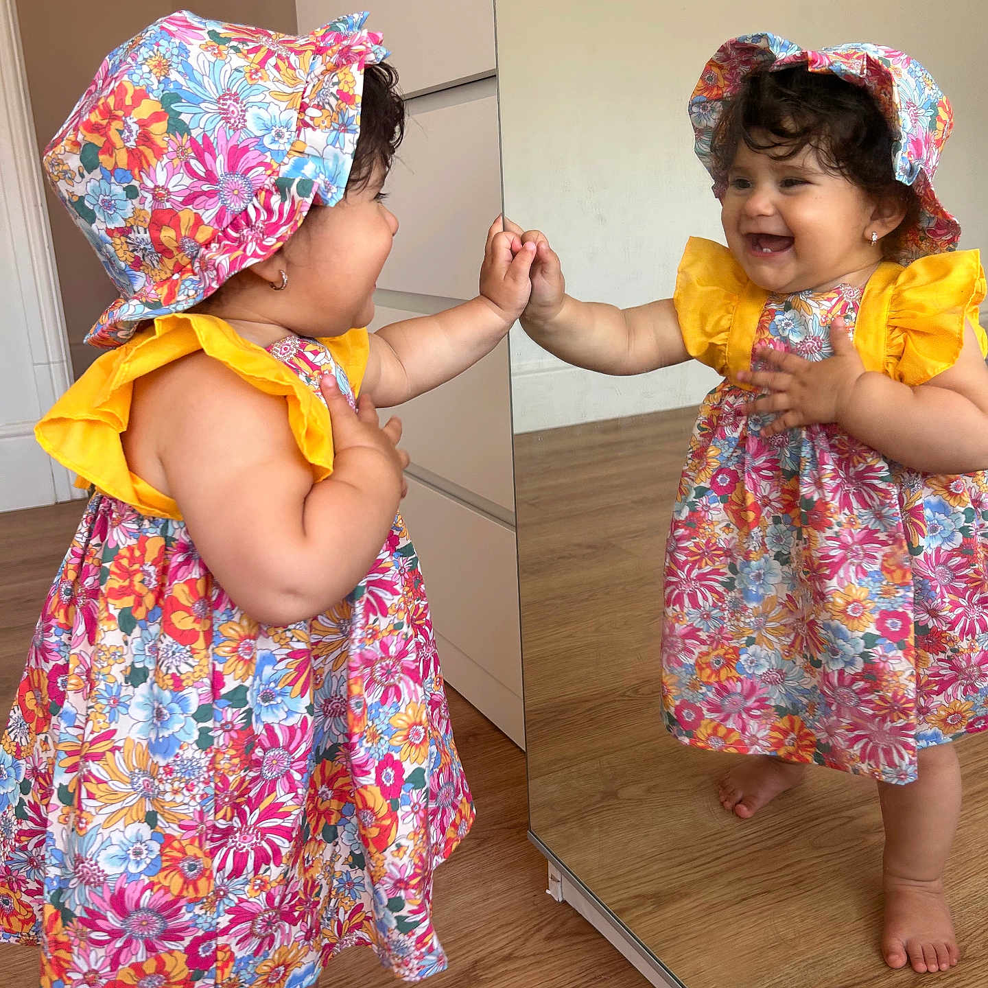 Mariam is registered to the contest to win money with this photo: barefoot, cabinet, child, colorful_clothing, curly_hair, cute, dress, earrings, floral_pattern, hand_touching, happy, hat, indoor, mirror, playful, reflection, smile, toddler, wooden_floor, young_child