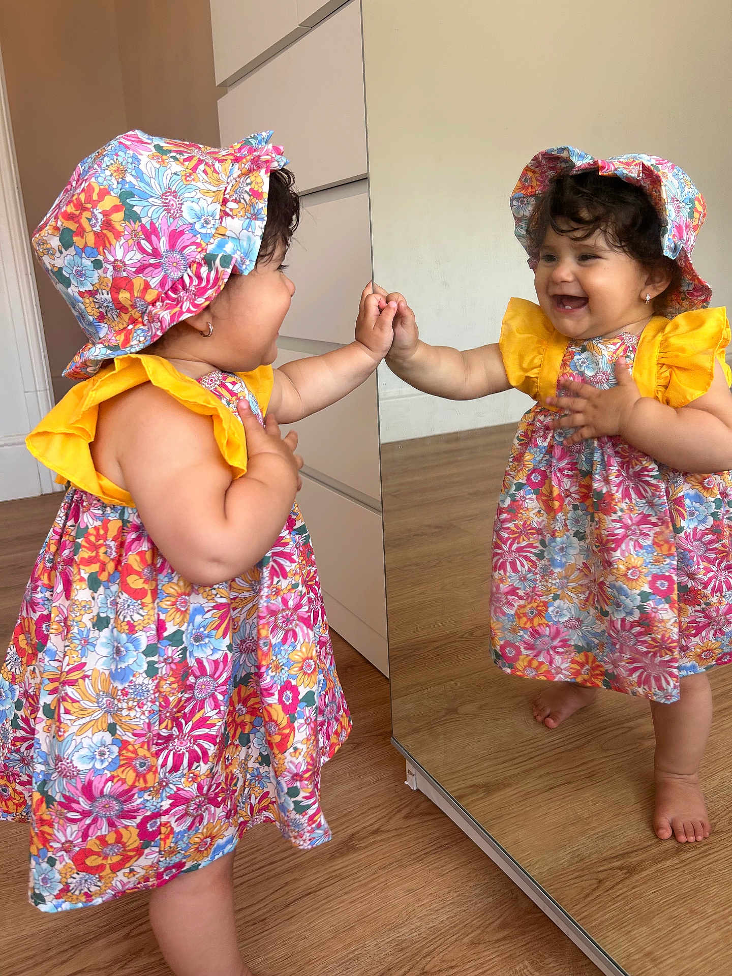 Mariam is registered to the contest to win money with this photo: toddler, child, mirror, reflection, dress, hat, floral_pattern, indoor, wooden_floor, barefoot, smile, happy, curly_hair, earrings, cabinet, hand_touching, colorful_clothing, young_child, playful, cute
