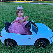 ángeles is registered to the contest to win money with this photo: toddler, child, pink_dress, bow, toy_car, white_car, grass, outdoor, happy, clapping, green_lawn, suburban, smiling, cute, play, vehicle, sunlight, park, small_car, dress_up