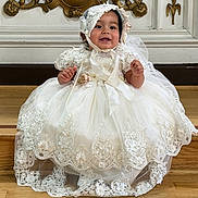 Aria is registered to the contest to win money with this photo: baby, child, infant, portrait, smiling, happy, sitting, white_dress, lace_dress, bonnet, formalwear, wedding_attire, ornate_background, gold_decoration, wooden_floor, indoor, home, photography, fashion, celebration