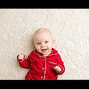 Tommy is registered to the contest to win money with this photo: baby, infant, red_pajamas, smiling, blue_eyes, lying_down, carpet, soft_texture, cute, happy, child, pajamas, indoors, portrait, one_person, light_skin, small_hands, buttoned_clothing, cozy, comfortable
