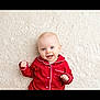 baby, infant, red_pajamas, smiling, blue_eyes, lying_down, carpet, soft_texture, cute, happy, child, pajamas, indoors, portrait, one_person, light_skin, small_hands, buttoned_clothing, cozy, comfortable