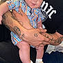 baby, child, infant, plaid_shirt, tattoos, adult, holding, slippers, socks, smiling, indoor, person, cute, happy, seated, blue_bedspread, black_clothing, closeup, family, love