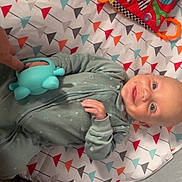 Hudson is registered to the contest to win money with this photo: baby, smiling, toy, hand, onesie, patterned_mat, colorful, indoor, infant, playtime, happy, cozy, child, person, blue_toy, fabric, cute, lying_down, human, interaction