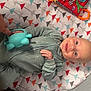 baby, smiling, toy, hand, onesie, patterned_mat, colorful, indoor, infant, playtime, happy, cozy, child, person, blue_toy, fabric, cute, lying_down, human, interaction