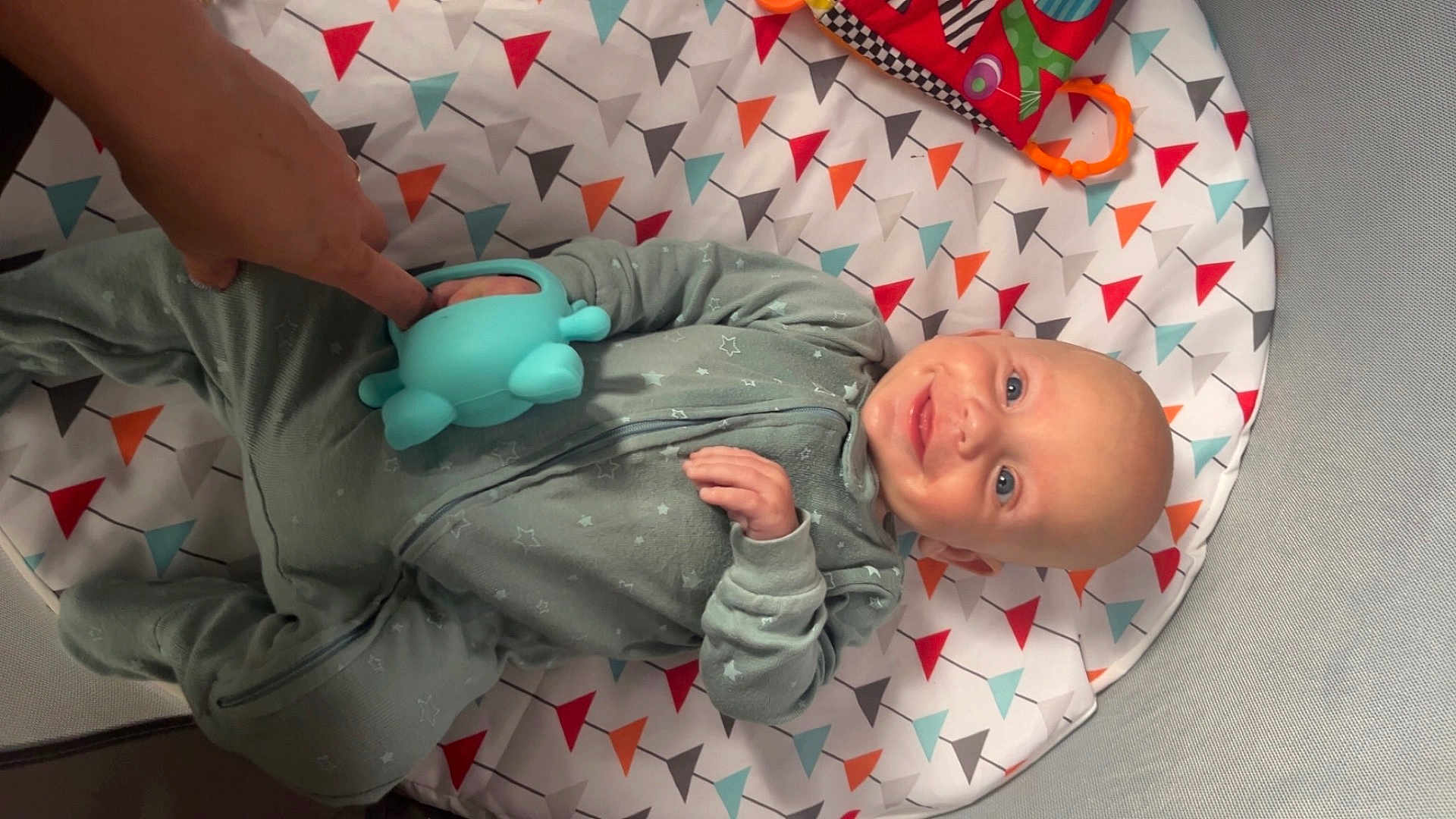 Hudson is registered to the contest to win money with this photo: baby, smiling, toy, hand, onesie, patterned_mat, colorful, indoor, infant, playtime, happy, cozy, child, person, blue_toy, fabric, cute, lying_down, human, interaction