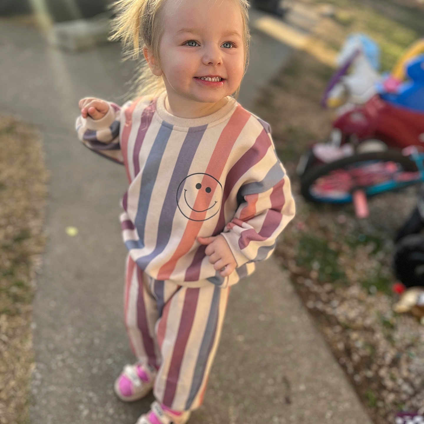 Korah joined the competition — help win amazing prizes! toddler, child, smiling, outdoor, sidewalk, sunlight, lens_flare, striped_clothing, topknot, toy, tricycle, happy, playful, casual_wear, footwear, person, daylight, young_child, hair_tie, background_blur