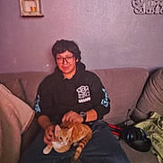 Huero is registered to the contest to win money with this photo: casual_clothing, cat, couch, cozy, decor, furniture, glasses, headphones, hoodie, indoor, living_room, nose_ring, orange_cat, person, petting, purple_wall, relaxed, shelf, sitting, watch