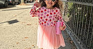 Aura is registered to the contest to win money with this photo: autumn, boots, buildings, child, daytime, fence, girl, heart_shaped, leaves, outdoor, pink_dress, portrait, sidewalk, smiling, strawberry_pattern, sunglasses, sunlight, tree, urban, water_bottle