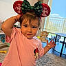 Arya is registered to the contest to win money with this photo: bracelet, carpet, child, clothing, cute, daylight, green_bow, headband, home, indoor, living_room, minnie_mouse, person, pink_shirt, playful, red_ears, smiling, toddler, toy, window
