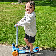 Jude joined the competition — help win amazing prizes! child, scooter, outdoor, grass, trees, sidewalk, smiling, happy, hoodie, shorts, shoes, sunny, daytime, play, park, person, young, boy, recreation, fun