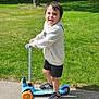child, scooter, outdoor, grass, trees, sidewalk, smiling, happy, hoodie, shorts, shoes, sunny, daytime, play, park, person, young, boy, recreation, fun