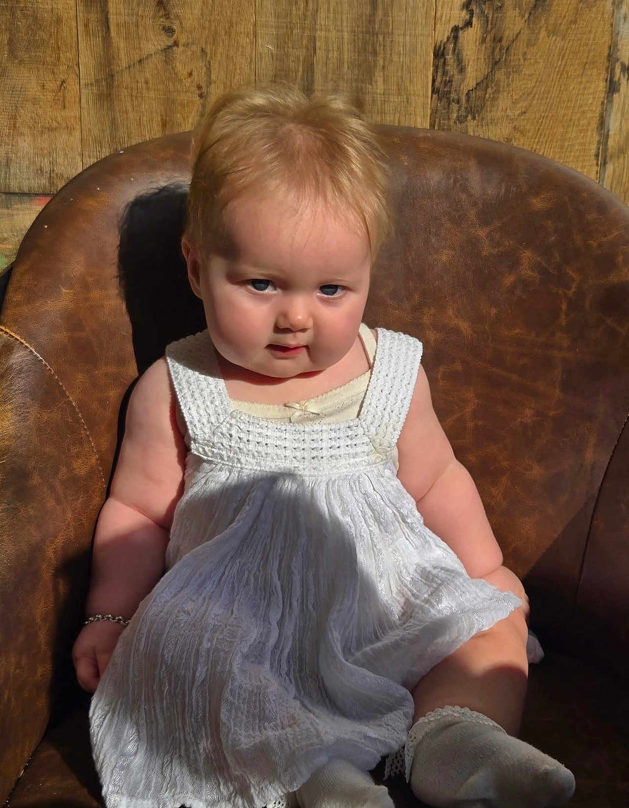 Amora-Louise is registered to the contest to win money with this photo: baby, child, white_dress, socks, leather_chair, wooden_background, sunlight, shadow, serious_expression, cute, indoors, portrait, infant, plush, bracelet, seated, face, skin, short_hair, candid