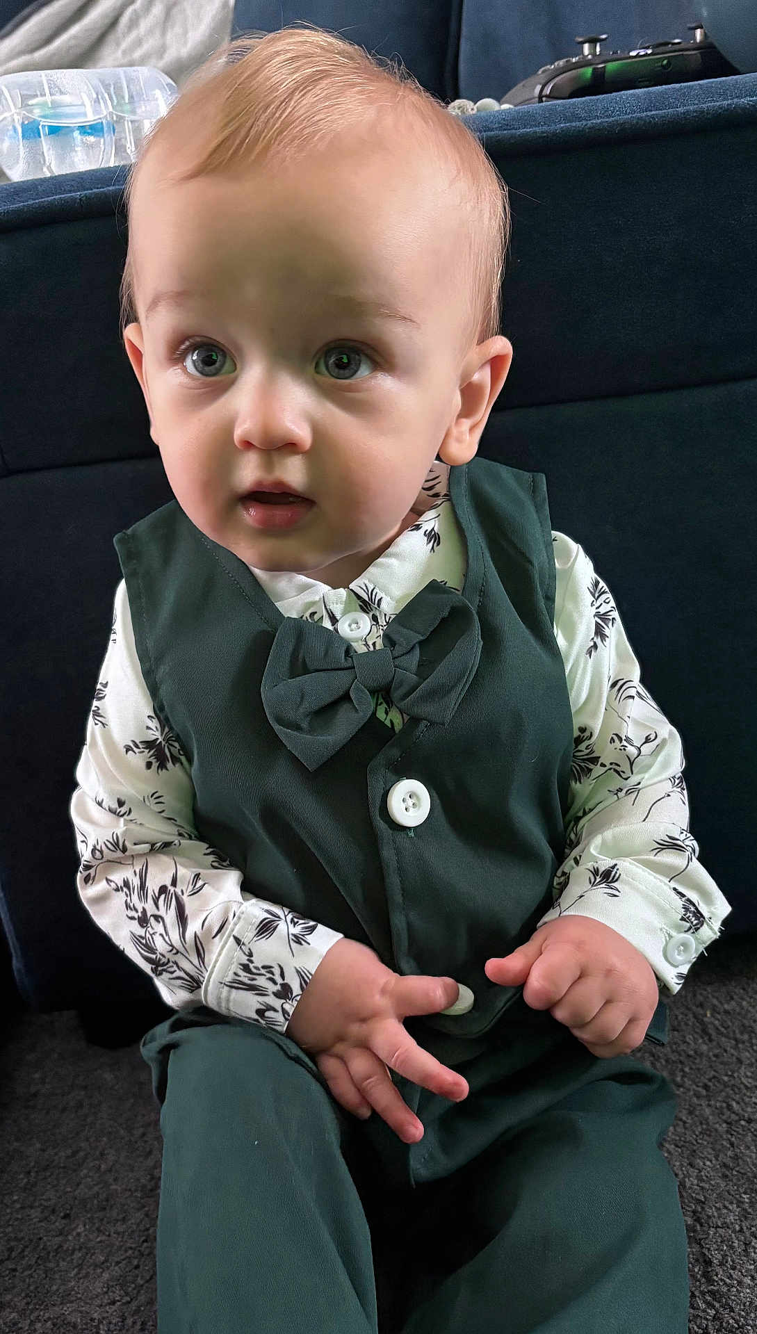 Reuben is registered to the contest to win money with this photo: toddler, child, boy, vest, bow_tie, shirt, green_clothing, sitting, carpet, indoor, curious_expression, hands, button, blond_hair, portrait, face, eyes, cute, young_child, fashion