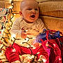 baby, blanket, celebration, child, christmas_lights, cozy, cute, decorations, festive, gift_bag, happy, holiday, holiday_season, indoors, red_pants, sitting, smiling, toddler, warm, white_shirt
