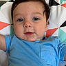 baby, infant, child, face, portrait, blue_shirt, brown_hair, blue_eyes, lying_down, blanket, geometric_pattern, bed, indoors, close_up, looking_at_camera, calm, drool, short_sleeves, colorful, photography