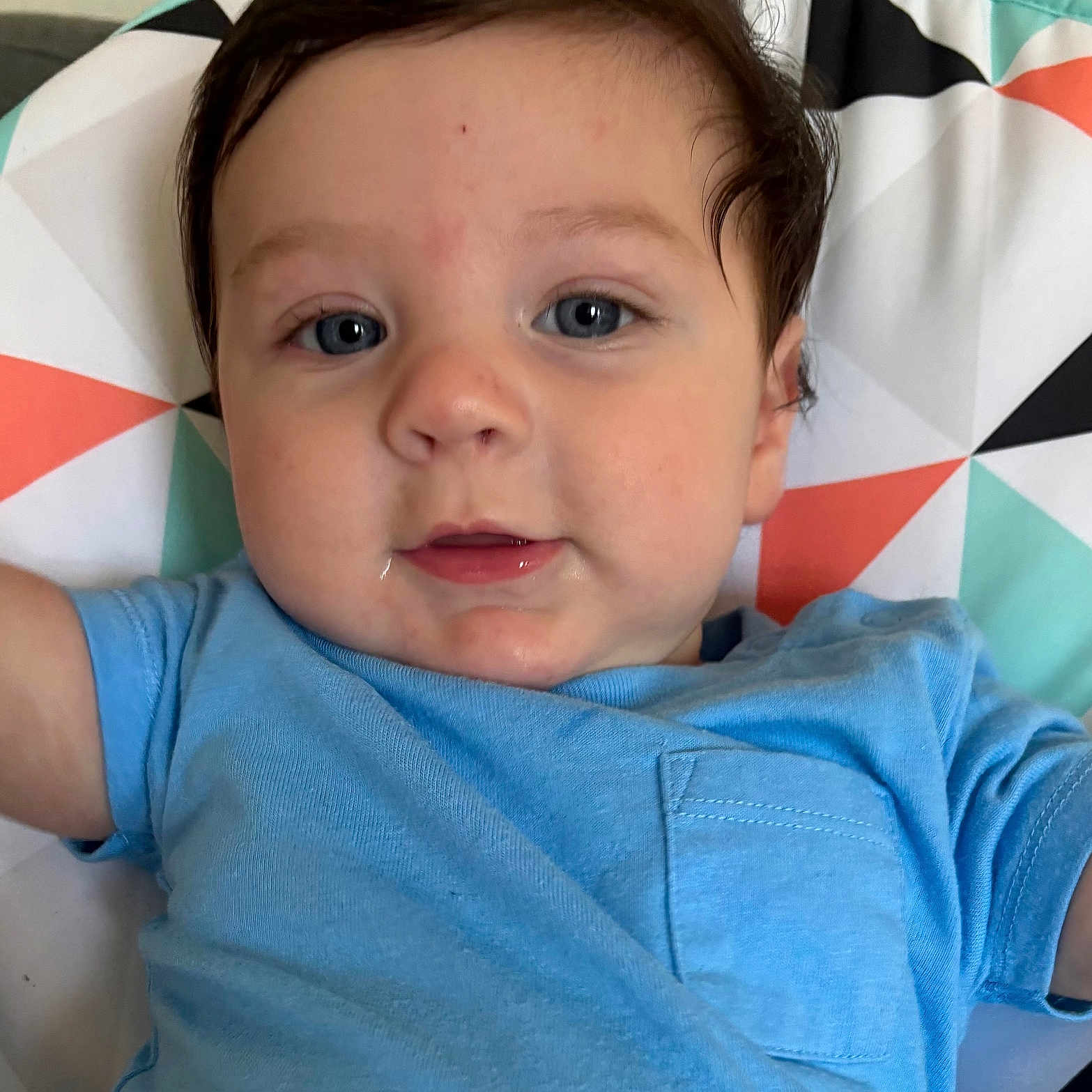 Jaylen joined the competition — help win amazing prizes! baby, infant, child, face, portrait, blue_shirt, brown_hair, blue_eyes, lying_down, blanket, geometric_pattern, bed, indoors, close_up, looking_at_camera, calm, drool, short_sleeves, colorful, photography