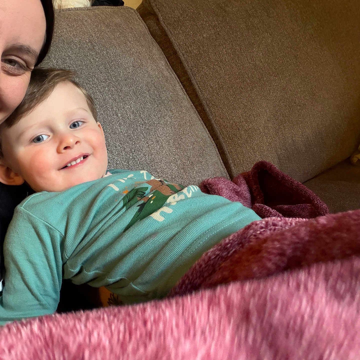 Ronnan is registered to the contest to win money with this photo: blanket, blue_shirt, casual_clothing, child, closeup, couch_cushion, cozy, face, family, hair, happy, indoor, living_room, parent_partial, portrait, relaxing, skin, smile, sofa, toddler