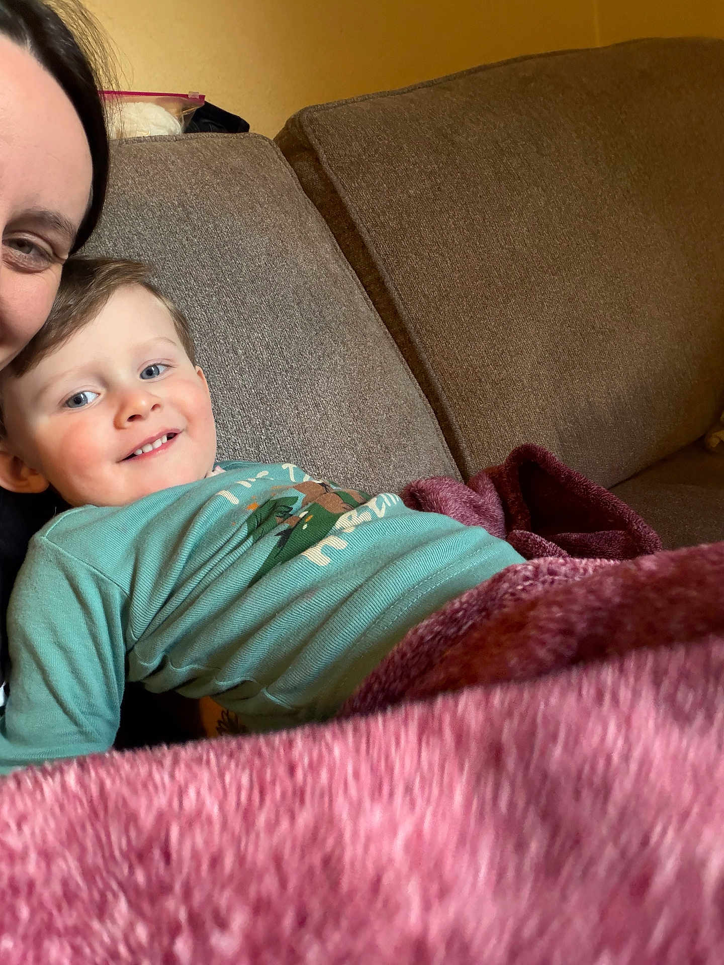 Ronnan is registered to the contest to win money with this photo: child, parent_partial, sofa, blanket, smile, toddler, blue_shirt, living_room, couch_cushion, portrait, face, happy, indoor, relaxing, closeup, family, hair, skin, casual_clothing, cozy